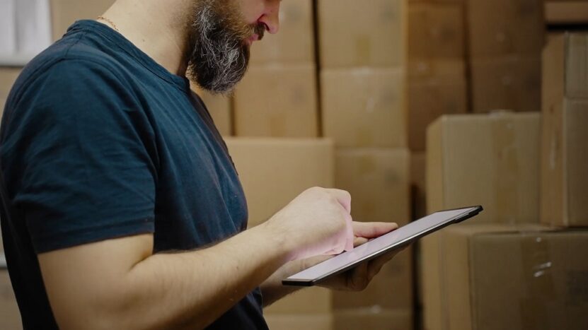 Warehouse worker checking inventory details on a tablet while standing among stacked shipping boxes