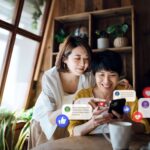 Happy Asian senior mother and daughter using smartphone together joyfully at home. Checking social media on mobile phone, receives notification, likes, views and comments. Social media