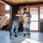 Two construction workers reviewing home renovation plans on a digital tablet inside an unfinished house.