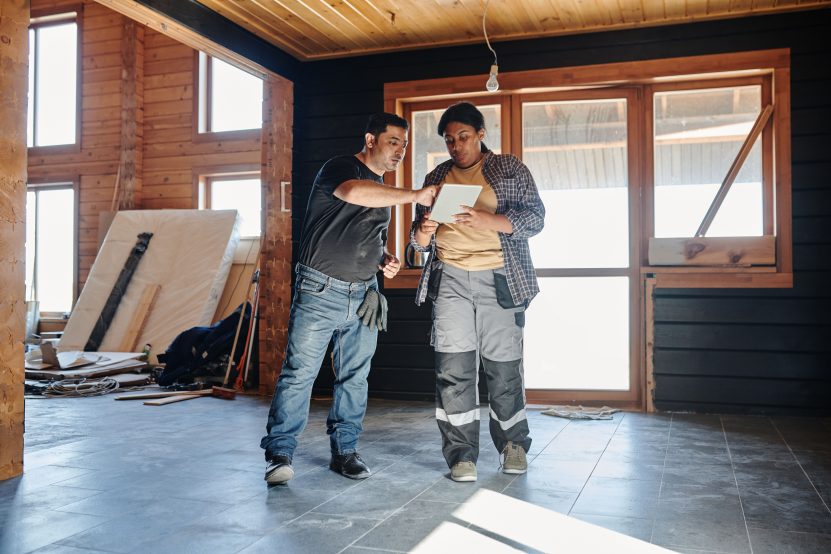 Two construction workers reviewing home renovation plans on a digital tablet inside an unfinished house.